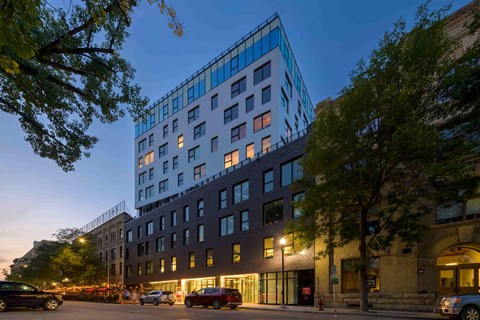 A modern building with a glass facade is illuminated from within at dusk.