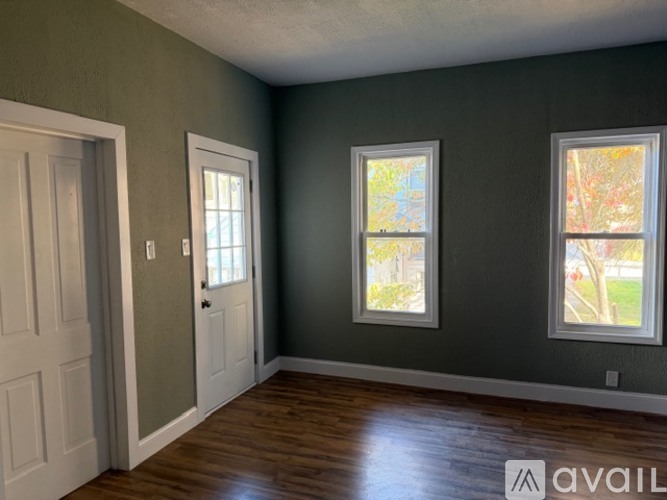 A room with a white door and two windows with a view of trees outside.