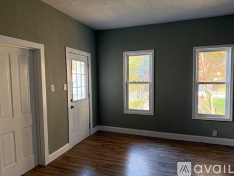 A room with a white door and two windows with a view of trees outside.