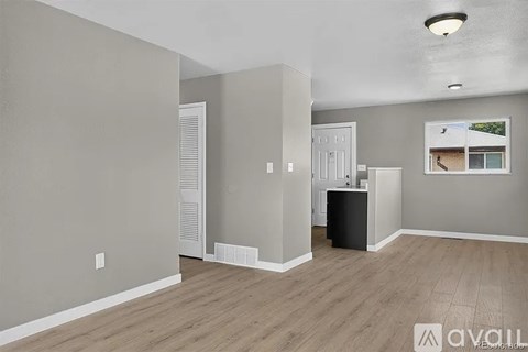 A spacious room with light grey walls and wooden flooring, featuring a white door, a window with a view of a house, and a black cabinet.