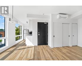 A spacious room with wooden flooring and white walls, featuring a black door and a kitchen area with white appliances.