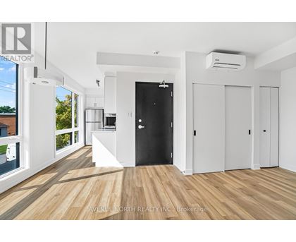 A spacious room with wooden flooring and white walls, featuring a black door and a kitchen area with white appliances.