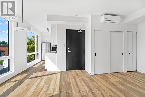 A spacious room with wooden flooring and white walls, featuring a black door and a kitchen area with white appliances.