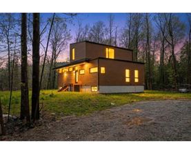 A modern house with a brown exterior is surrounded by trees and has a gravel driveway leading to the front door.