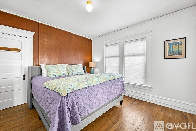 A bedroom with a bed covered in a purple comforter and a painting on the wall.