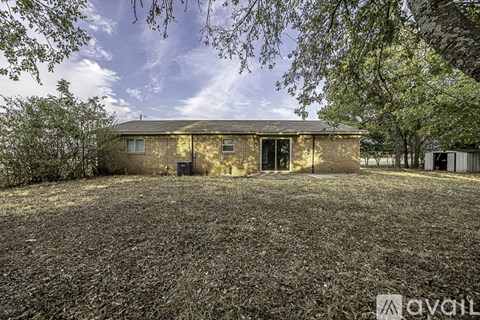A house with a brown brick exterior is surrounded by trees and has a gravel driveway in front.