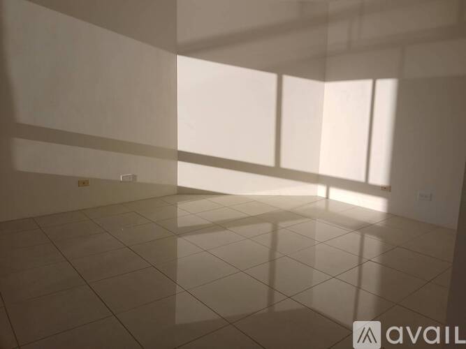 A spacious room with white tiled flooring and large windows allowing natural light to enter.