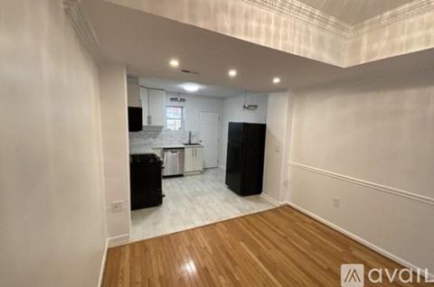 A room with wooden flooring and white walls, with a kitchen area visible in the background.