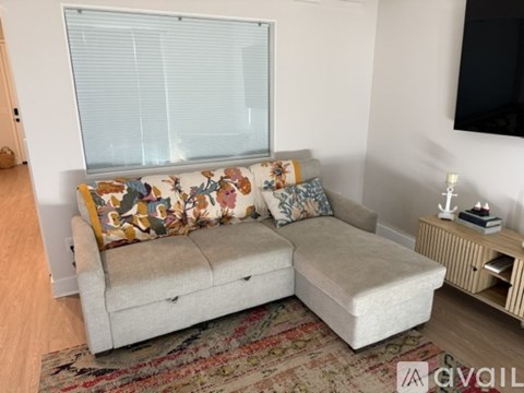 A couch with a floral patterned pillow is in the foreground of a room with a window and a wooden floor.