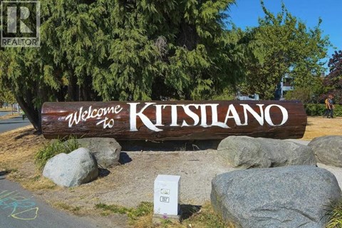 A wooden sign that says "Welcome to Kitsilano" is in front of some trees.