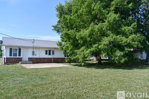 A house with a white exterior and a grey roof is surrounded by a grassy lawn and a large tree.