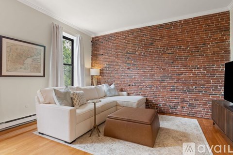 A living room with a white couch and a brown ottoman in front of a brick wall.