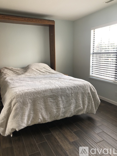 A bed with a white comforter is in a room with wooden floors and a window with blinds.