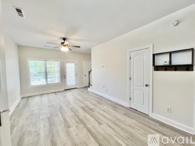 A spacious room with a ceiling fan and light, hardwood flooring, and a large window with blinds.
