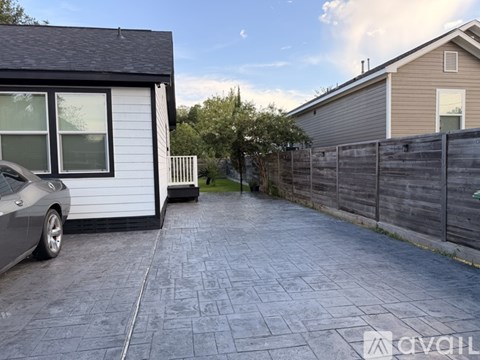 A grey car is parked in a driveway with a white house and a wooden fence.