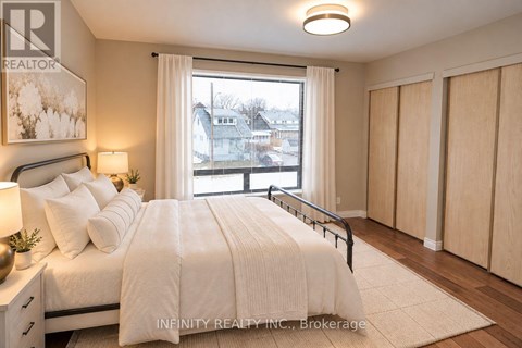 A bedroom with a large bed, a nightstand with a lamp, and a window with a view of the neighborhood.