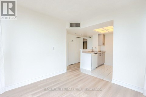 A white kitchen with a sink and cabinets is seen in a room with white walls and wooden flooring.