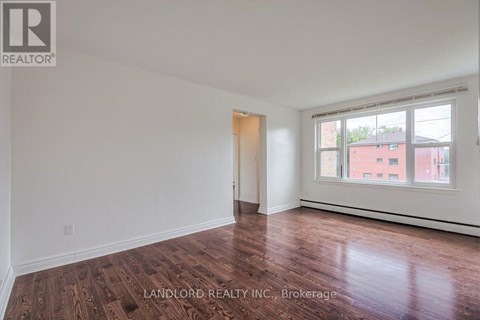 A room with wooden flooring and white walls, with a window showing a red building outside.