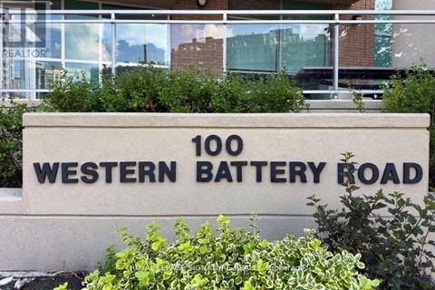 A sign that says "100 Western Battery Road" in front of some bushes.