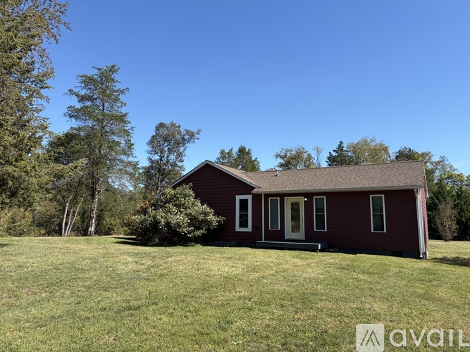 A small house with a brown roof and white trim is surrounded by a grassy field and trees.