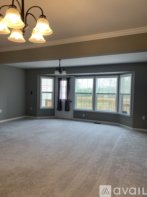 A room with a carpeted floor, a chandelier, and a large window with a view of a fence and trees.