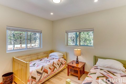 A bedroom with two beds, one with a quilt and the other with a blanket, a small wooden table with a lamp, and windows with blinds.