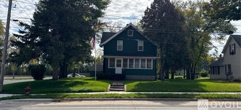 A green house with white trim is situated on a street with a tree in front of it.