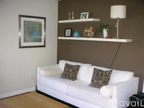 A white couch with two brown pillows sits in front of a wall with a framed picture and a shelf with various decorative items.