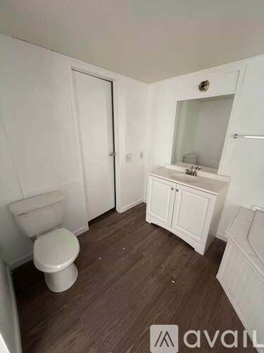 A white toilet sits in a small bathroom with wooden floors and white walls.