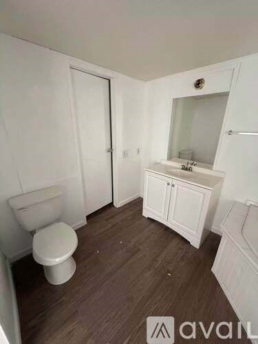 A white toilet sits in a small bathroom with wooden floors and white walls.