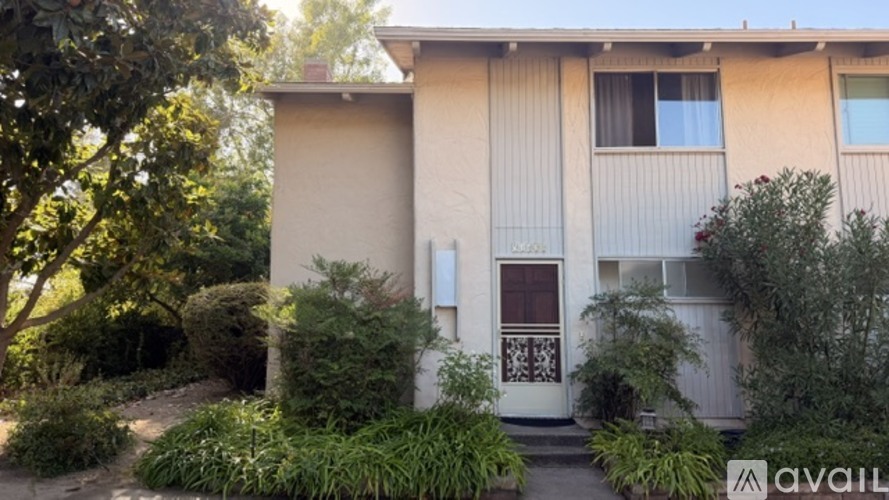 A beige house with a white door and windows surrounded by greenery.
