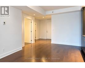 A room with white walls and wooden flooring, with a door on the left and a kitchen area on the right.