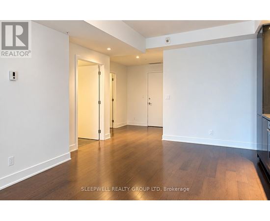 A room with white walls and wooden flooring, with a door on the left and a kitchen area on the right.