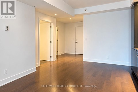 A room with white walls and wooden flooring, with a door on the left and a kitchen area on the right.