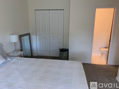 A bedroom with a bed, a nightstand with a lamp, a mirror, a trash can, and a toilet in the hallway.