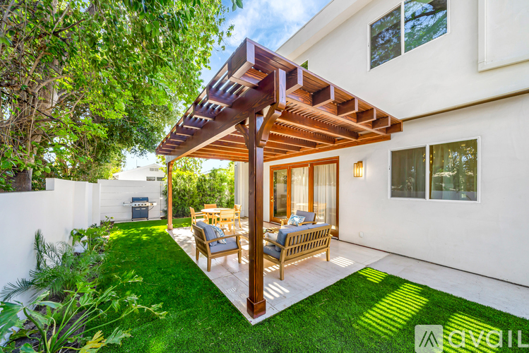 A patio with a wooden pergola and chairs is surrounded by a white wall and greenery.