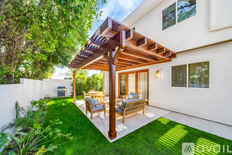 A patio with a wooden pergola and chairs is surrounded by a white wall and greenery.