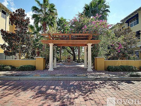 A wooden archway leads to a building with a brick pathway and palm trees.