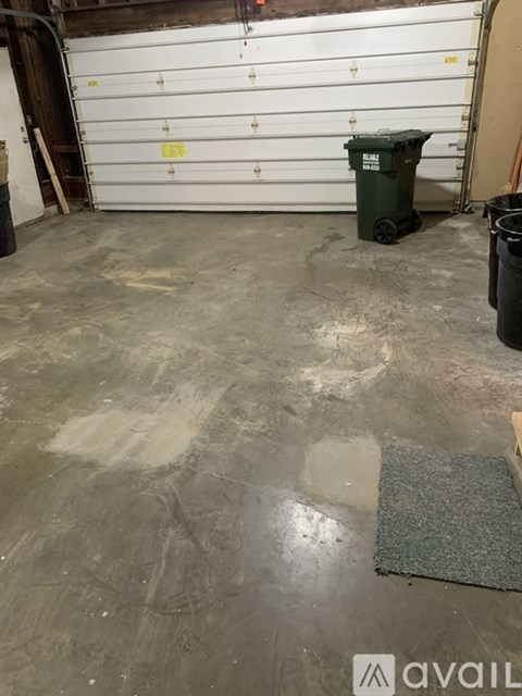 A dirty garage floor with a green trash can and a grey mat.