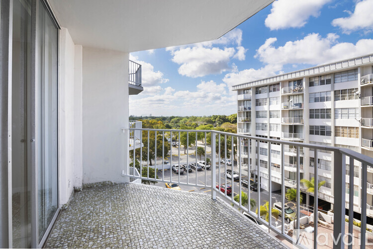 A balcony with a metal railing and a tiled floor overlooks a parking lot and buildings.