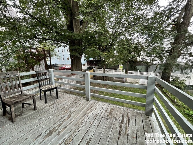 Two chairs are on a wooden deck with a fence and trees in the background.