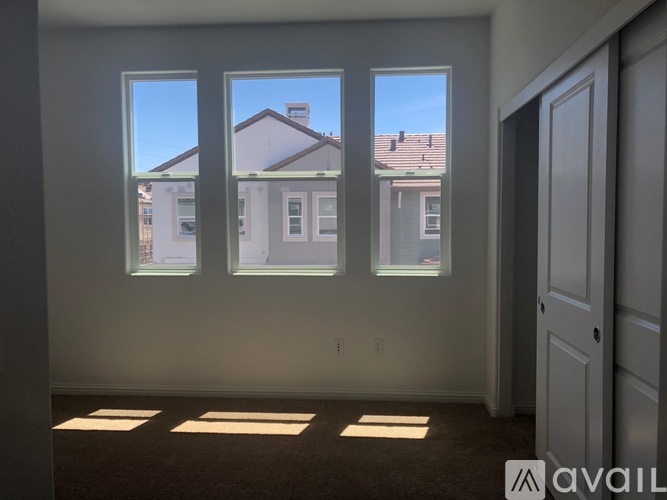 A room with a carpeted floor and three windows overlooking houses.