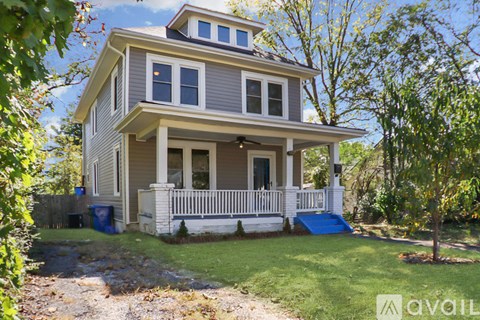 A house with a grey and white exterior is surrounded by trees and has a blue mat on the front porch.