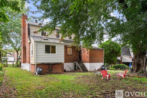 A house with a red brick chimney and a white siding is surrounded by trees and has a patio with red chairs.