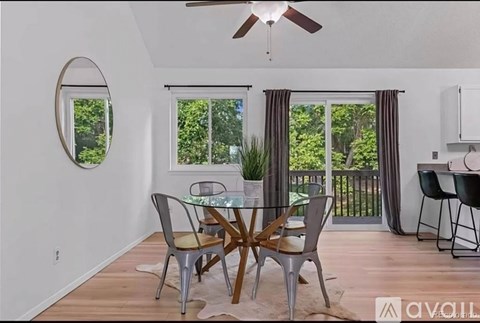 A dining room with a glass table and chairs, a ceiling fan, and a view of trees through the window.