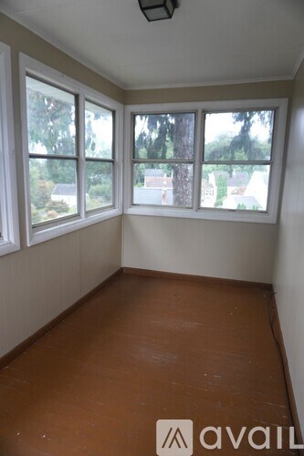 A room with brown floors and white walls has a window with a view of trees and houses.
