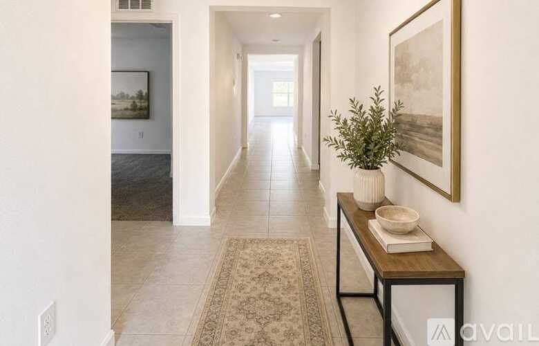 A hallway with a rug, a table with a bowl and a plant on it, and a painting on the wall.