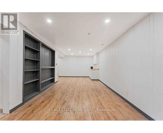 A spacious room with wooden flooring and white walls, featuring a row of shelves on the left side.