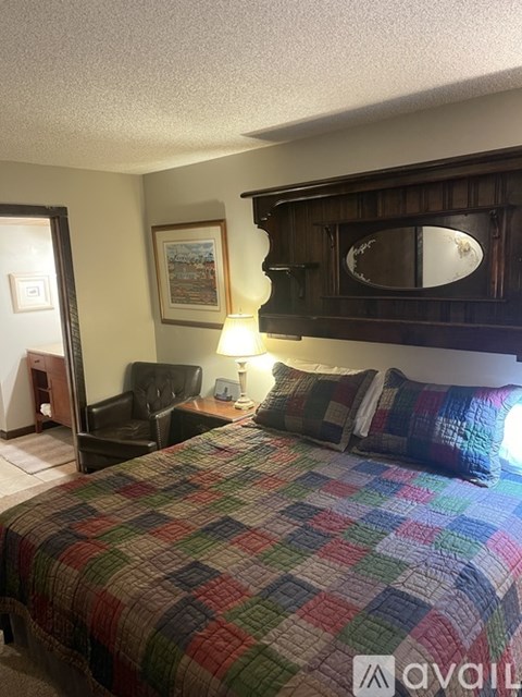 A bedroom with a bed covered in a multicolored quilt, a chair, and a lamp.