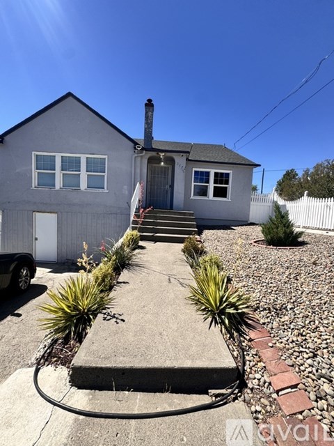 A house with a grey exterior and a white door is surrounded by a gravel area and some green plants.